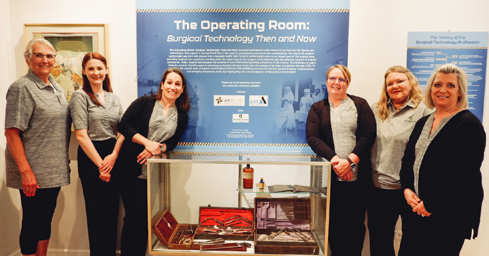 Six smiling women standing around Surgical Technology museum exhibit