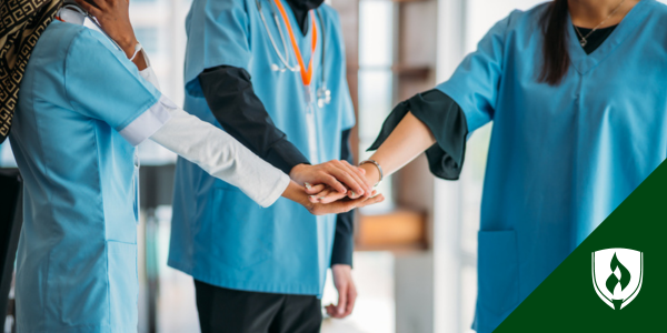 Three nurses in blue scrubs put their hands together in a circle